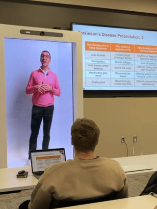 holographic box displaying a man lecturing to a class with a presentation to the side of him